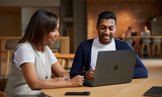 Amar colaborando con un colega mientras está sentado con su MacBook en la mesa de un área común.
