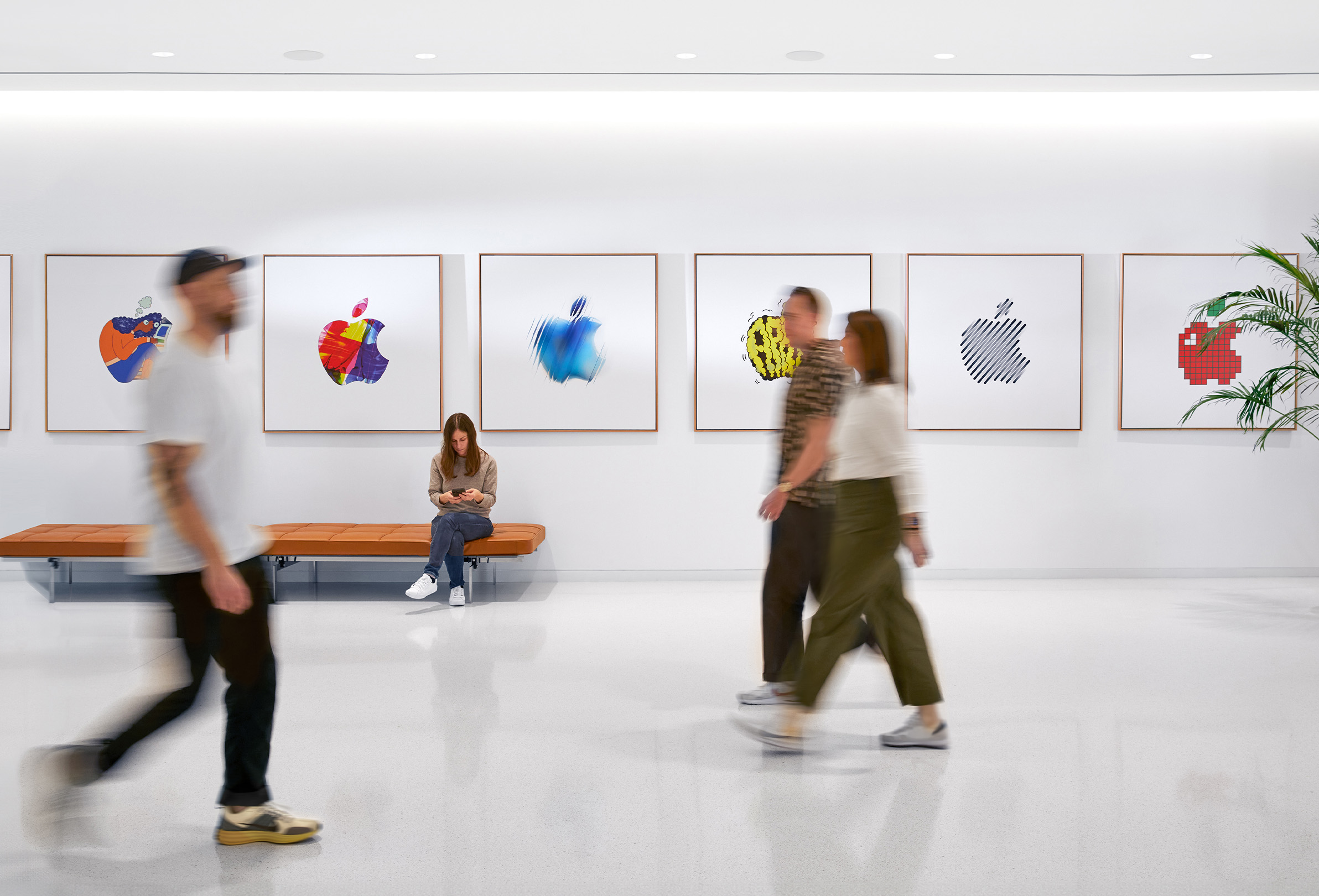 Tre dipendenti Apple camminano in uno spazio interno davanti a un muro con loghi Apple colorati; su una panca è seduta una quarta persona.