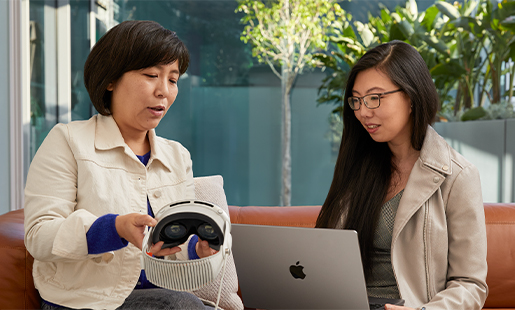 Jessica che usa un MacBook ed è seduta insieme a una collega Apple che le sta mostrando Apple Vision Pro; dalla porta si intravede il patio esterno.