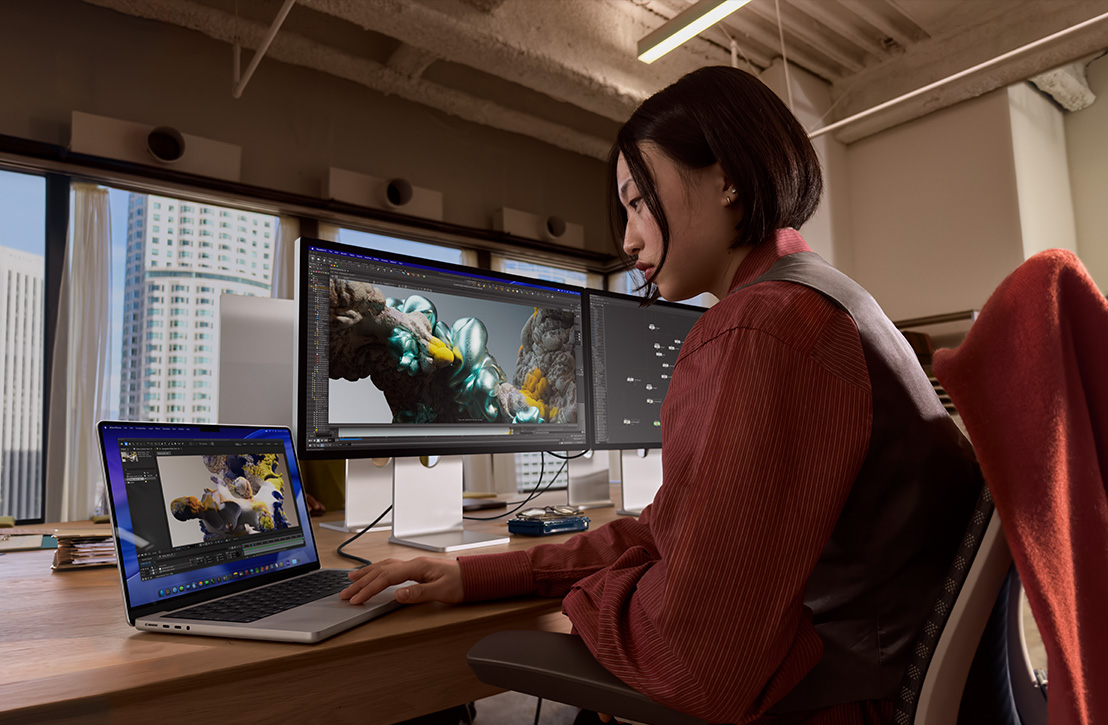 Seseorang bekerja di meja menggunakan MacBook Pro yang terhubung ke layar eksternal, kedua layar menampilkan rendering 3D, sementara gedung-gedung terlihat melalui jendela di belakangnya