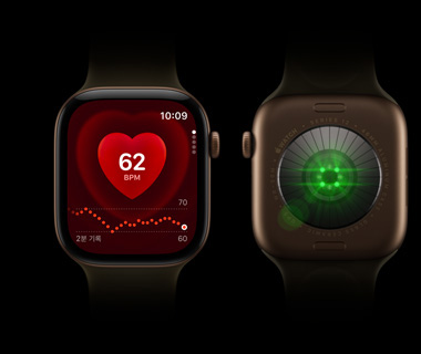 나란히 놓인 Apple Watch Series 12 기기 두 대. 왼쪽에는 Apple Watch Series 앞모습에 심박수 모니터가 표시되어 있고, 오른쪽에는 Apple Watch Series 12의 뒷모습이 보이고, 초록색으로 밝게 빛나는 빛이 보입니다.