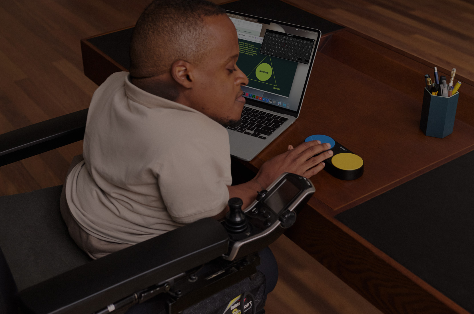 Photo of a man in a power wheelchair at a desk, using Accessibility Keyboard feature on his 13-inch MacBook Air and a switch control device