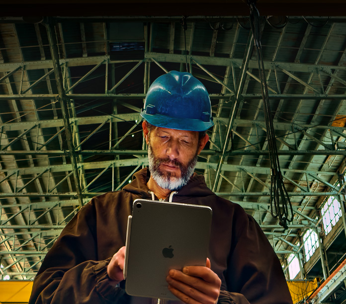 iPad, color plata, con un Apple Pencil adherido, sostenido por una persona con chamarra café y casco azul, trabajando en un entorno industrial con vigas de metal