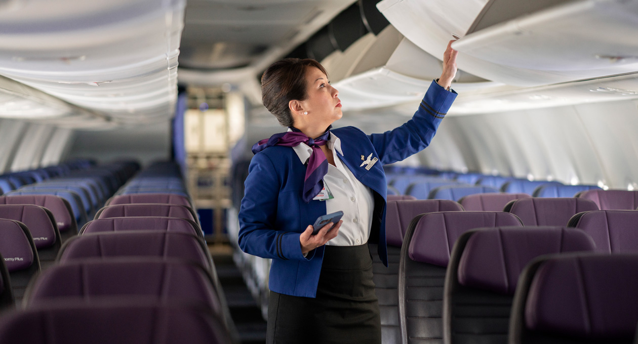 Una azafata sujeta un iPhone mientras abre un compartimento superior en un avión vacío