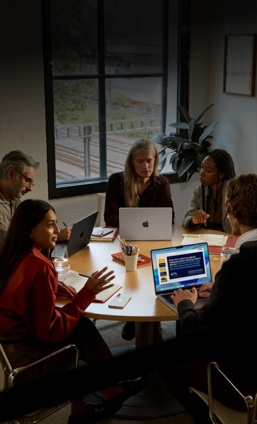 Five individuals seated around a conference table, collaborating on MacBooks, with iPhones face down on the table