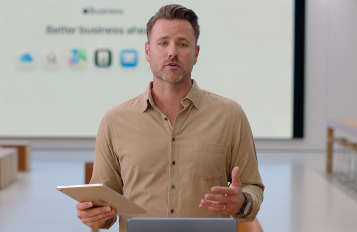 Presenter speaks while referencing an iPad, and a large presentation screen displays various business app icons.