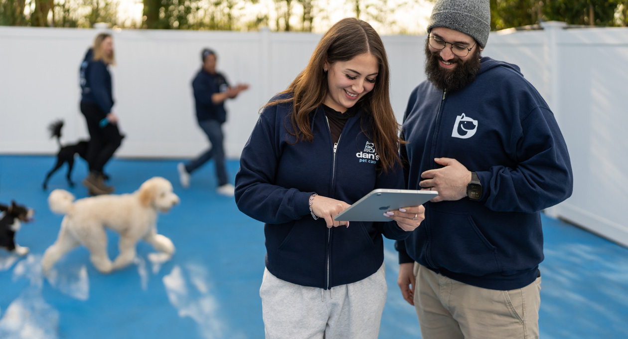 Two employees in matching branded hoodies look at an iPad together in a dog play area, dogs and staff visible in the background