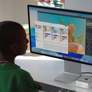 Uma pessoa sentada trabalha em uma planilha do Excel intitulada "Acompanhamento de vendas do quarto trimestre" no iMac e no Mac mini. Atrás da planilha do Excel está uma janela do Pixelmator Pro e o fundo do macOS Tahoe