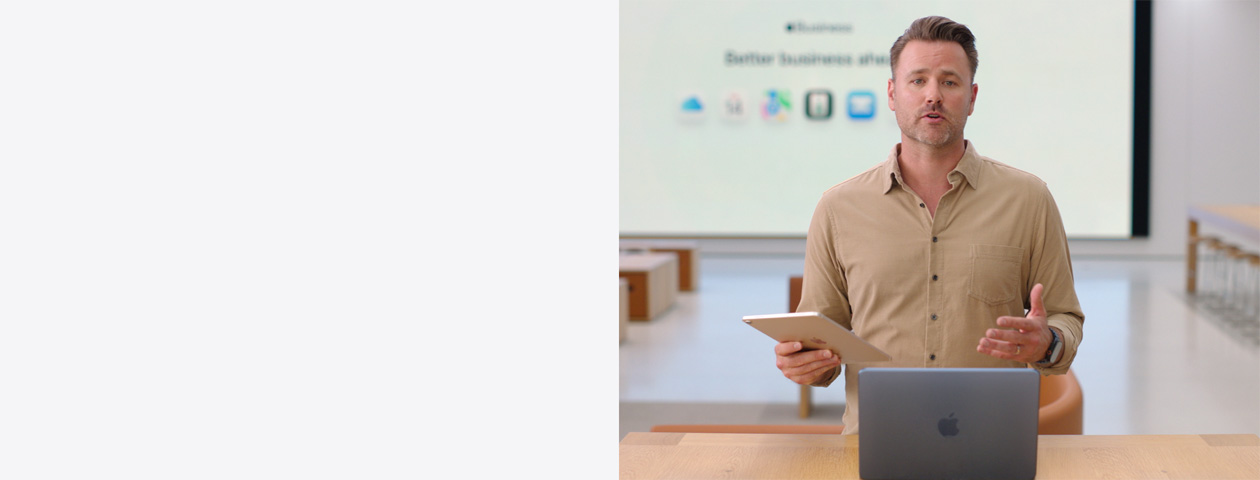 Un presentatore parla tenendo in mano un iPad di fronte a un MacBook aperto su un tavolo; alle sue spalle un grande schermo mostre le icone di alcune app per il lavoro.