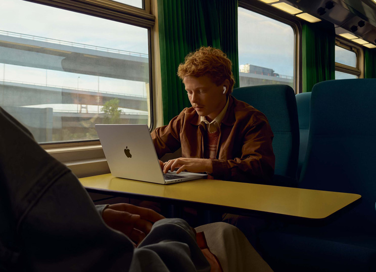 Junassa ikkunapaikalla istuva henkilö käyttää AirPodeja ja työskentelee MacBookilla.