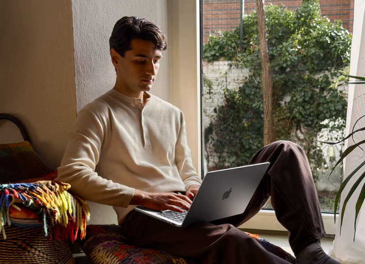 Una persona sentada en el suelo junto a una ventana está usando un MacBook.