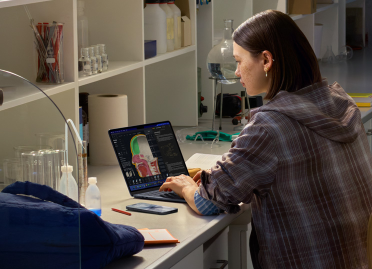Una persona sentada en un laboratorio usa un MacBook; la pantalla muestra varias apps.