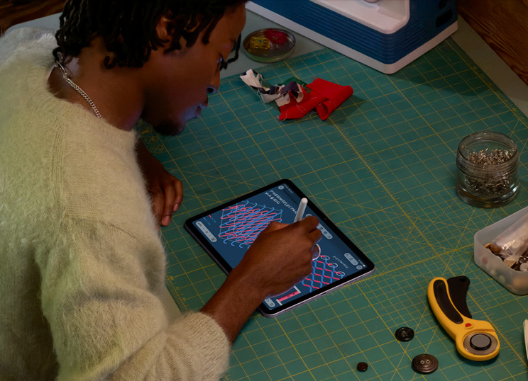 Una persona dibuja con el Apple Pencil en un iPad, apoyada sobre una base de corte verde con materiales de costura.