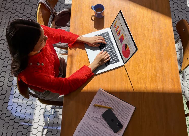 Una persona usa un MacBook en una mesa, con una taza, un smartphone y papeles cerca.