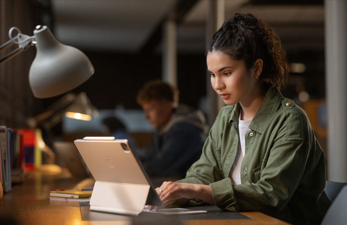 A student sits at a desk in a study space, typing on an iPad with Magic Keyboard, demonstrating versatility of Apple devices