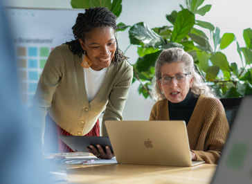 Two professors collaborate together at a desk, one holds an iPad while the other works on Mac