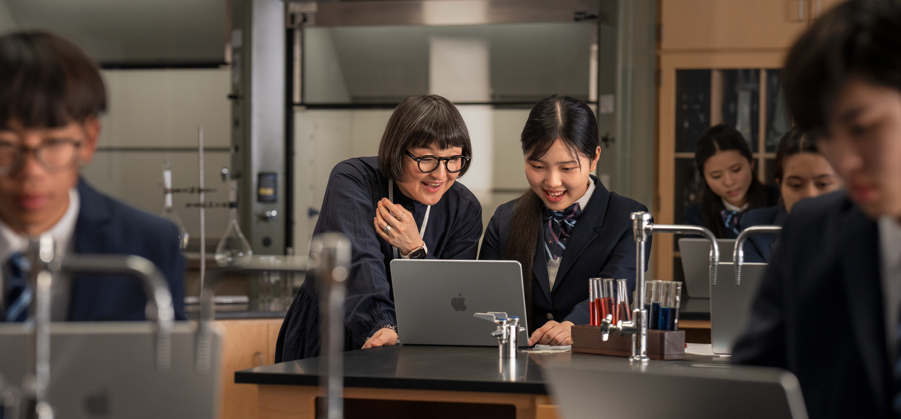 實驗室環境中，桌上放著打開的 MacBook，學生與教師在旁互動，周圍擺著試管和設備