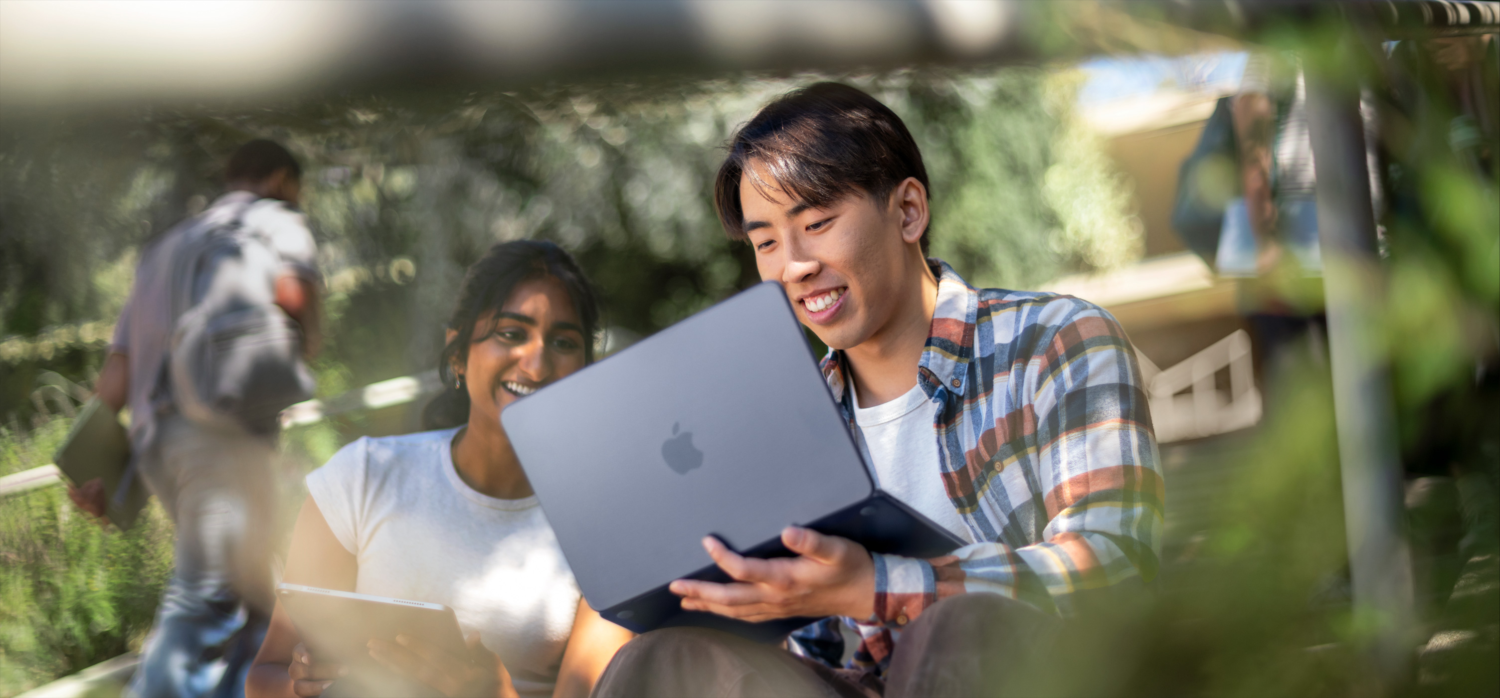 Two smiling students use MacBook outdoors, one holds it on their lap as they collaborate on an assignment