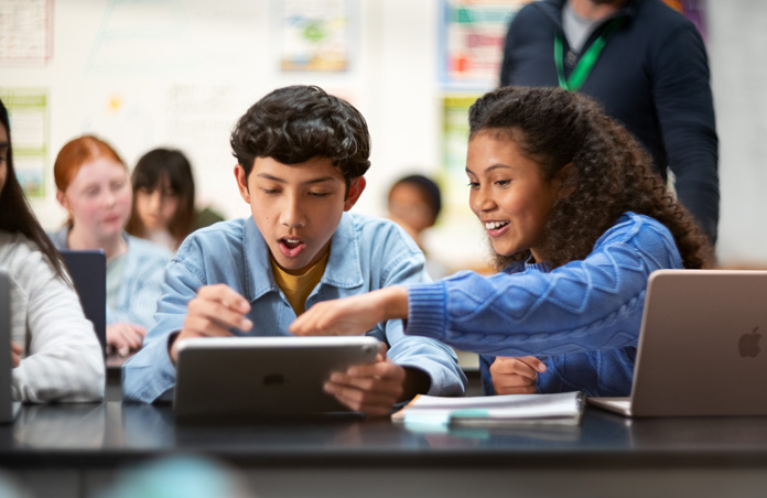 Two students point to iPad screen in excitement, while a MacBook is also open on the desk