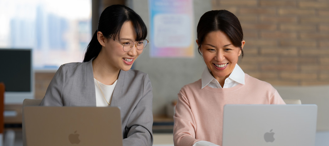 Two smiling educators sit together collaborating at a desk, each with a MacBook open