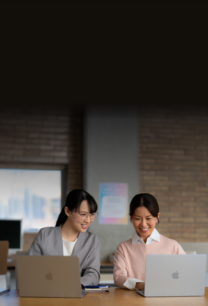 Two smiling educators sit together collaborating at a desk, each with a MacBook open