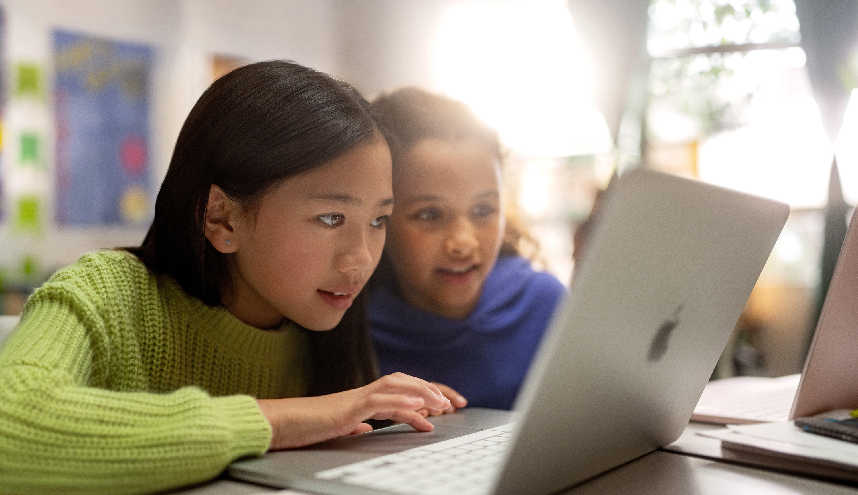 Two young students seated at a desk looking intently at MacBook as they collaborate in a classroom