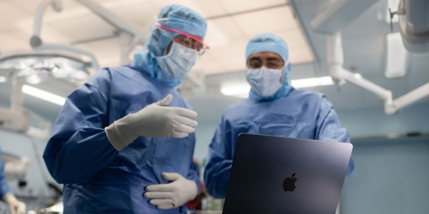 Två kirurger står i en operationssal och tittar på en MacBook. Den ena gestikulerar med handen medan den andra ser på.