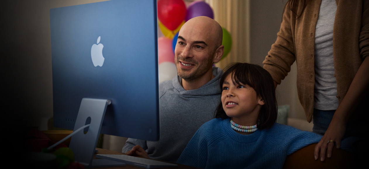 Two people sitting together use an iMac while a third person stands behind them observing.