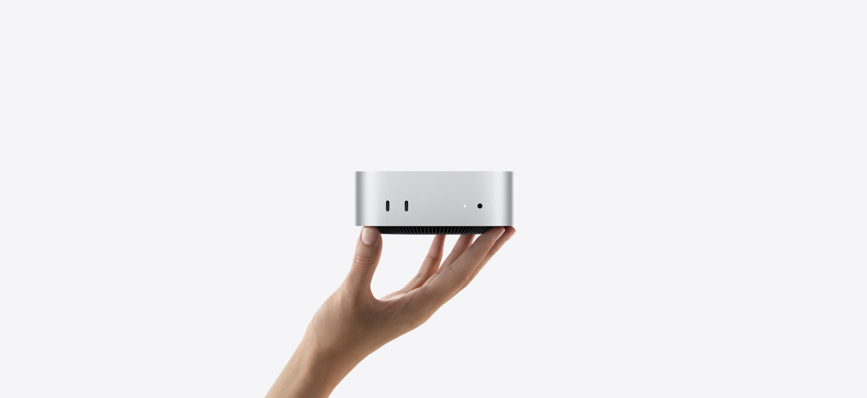 Mac mini zepředu na prstech natažené ruky, dva porty Thunderbolt, stavová kontrolka a sluchátkový konektor na přední straně, černá základna se zkosenými hranami na spodní straně, plochá horní strana, zaoblené boční hrany, rovné strany, stříbrná barva