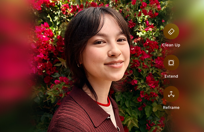 Photo of smiling person with brown hair, wearing a brown sweater, surrounded by red flowers, AI photo editing demo with Clean Up, Extend, Reframe options shown over photo
