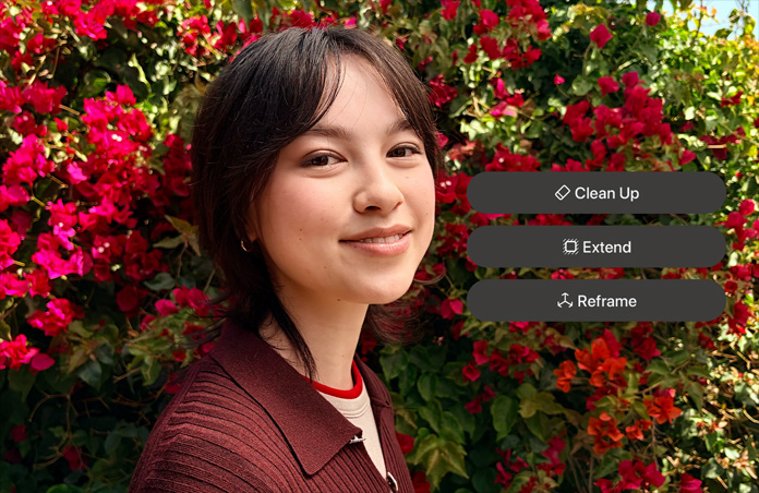 Photo of smiling person with brown hair, wearing a brown sweater, surrounded by red flowers, AI photo editing demo with Clean Up, Extend, Reframe options shown over photo